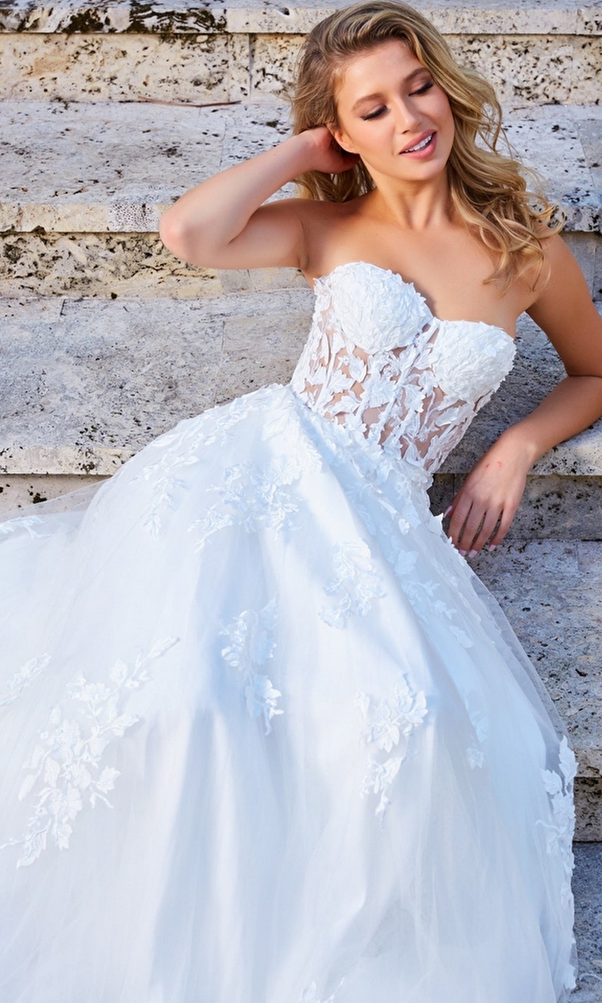 A woman wearing the Jovani Strapless Long Lace A-Line Prom Dress 07901 with floral details sits on stone steps, looking to the side and smiling.